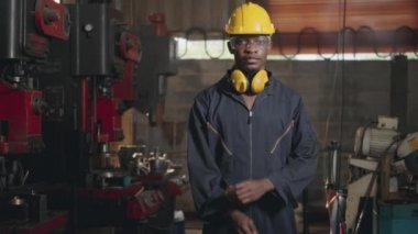 Makinenin önünde sarı başlık ve kulaklıkla gülümseyen Amerikalı endüstriyel işçi portresi endüstri fabrikasında kollarını kavuşturmuş mutlu mühendis. yavaş çekim