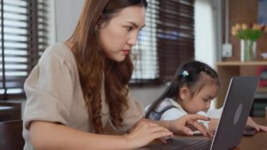 Asyalı genç anne evde dizüstü bilgisayarla çalışıyor ve kızının ev ödevlerini yapmasına yardım ediyor, bekar anne evden çalışıyor ve kızına masa başında ödev yaparken yardım ediyor.