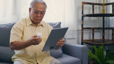 Dijital tabletle Asyalı yaşlı hastaların evdeki oturma odasında hangi ilaç şişesinin alınması gerektiğini öğrenmek için doktorlara video çağrısı, kıdemli yaşlı adam teknolojisi çevrimiçi sağlık hizmeti
