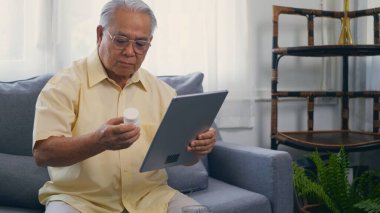 Asian elderly patient video call by digital tablet to doctor for inquire about which bottle of medication pills should be taken in living room at home,  Senior old man technology online healthcare clipart