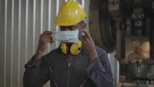 肖像画アメリカの工業用黒の若い労働者の男は フロントマシンで黄色のヘルメットと耳の保護と笑みを浮かべて 産業工場で働くに立って顔マスクを身に着けている幸せなエンジニア — ストック動画