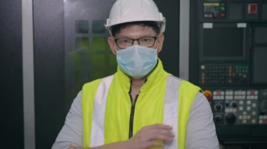 Asyalı fabrika işçisi, teknisyen ya da mühendis koruyucu maske ve miğfer takıyor. Endüstriyel fabrikada makinenin önünde kollarını kavuşturup kameraya bakıyor.