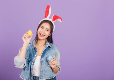 Mutlu Paskalyalar. Tavşan kulağı takmış, gülümseyen güzel bir genç kadın ellerinde renkli Paskalya yumurtaları, kameraya bakan bir kadın portresi, mor arka planda izole edilmiş bir stüdyo resmi.