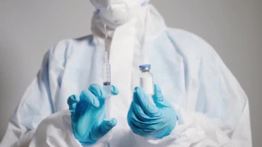 PPE süit üniformalı kadın doktor ya da bilim adamının yakından incelediği mavi eldiven giyen laboratuvar koruyucu ilaç şişesi, şırınga, koronavirüs ya da COVID 19