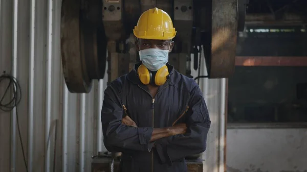 Portrait American industrial black young worker man smiling with yellow helmet and ear protection in front machine, Happy engineer remove mask from face at work in industry factory.