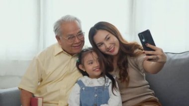 Mutlu aile zamanları, rahatla. Asyalı büyükanne ve büyükbaba, torunlarıyla cep telefonuyla evdeki kanepede selfie çekerken gülüyorlar. Büyük adam ya da büyükbaba, kız ve kız birlikte oturma odasında.