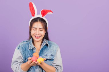 Mutlu Paskalyalar. Tavşan kulağı takmış, gülümseyen güzel bir genç kadın ellerinde renkli Paskalya yumurtaları, yumurtalara bakan bir kadın portresi, mor arka planda izole edilmiş bir stüdyo resmi.