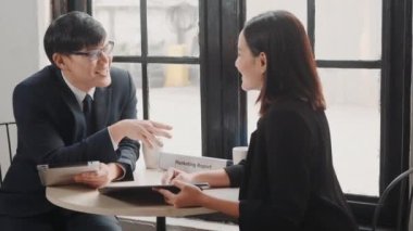 Mutlu iki Asyalı genç işadamı ve kadın buluşmadan önce el sıkışıyor veya dijital tabletle masaya oturuyor, iş adamlarının planlarını tartışıyor ve gülümsüyor.