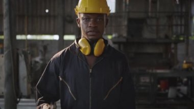 Ön makinede miğfer ve kulak koruyucusuyla gülümseyen Amerikalı endüstriyel işçi, fabrikada çalışırken omzunda ingiliz anahtarı tutan mühendis..