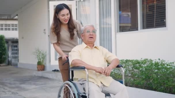 車いすの上の障害のあるシニア男性と娘 幸せなアジアの世代の家族が一緒に屋外の裏庭を楽しんで ケアヘルパー若い女性が高齢者の男を歩いて笑顔と笑い スローモーション — ストック動画