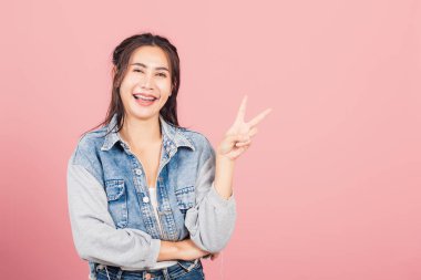 Asyalı mutlu bir portre güzel güzel bir kadın kot pantolon giy gülümse parmağınla zafer işareti işareti işareti yap uzağa bak kamera stüdyosuna bak pembe arka planda izole edilmiş fotokopi alanı ile