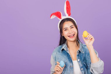 Mutlu Paskalyalar. Tavşan kulağı takmış, gülümseyen güzel bir genç kadın ellerinde renkli Paskalya yumurtaları, kameraya bakan bir kadın portresi, mor arka planda izole edilmiş bir stüdyo resmi.