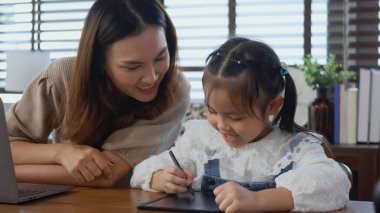 Asyalı genç anne evde dizüstü bilgisayarla çalışıyor ve kızının ev ödevlerini yapmasına yardım ediyor, bekar anne evden çalışıyor ve kızına masa başında ödev yaparken yardım ediyor.