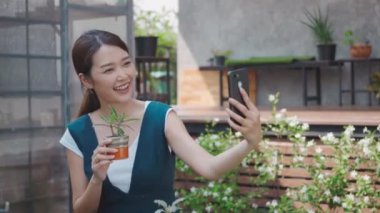 Mutlu Asyalı güzel genç bir kadın sera yakınlarındaki bir bahçede akıllı telefondan küçük bir ağacın selfie fotoğrafını çekiyor. Kadın bahçıvan videosu dijital cep telefonundan canlı yayında, ev bitkisinden.