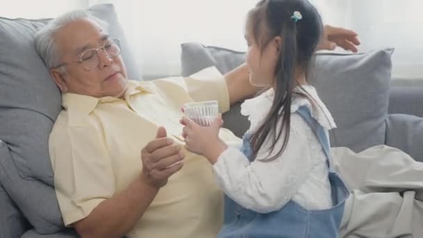 アジアの孫娘は おじいちゃんがリビングルームのソファで食べるために水のガラスを持ってきました Happy Little Kid Girl Smiling Giving Wather Old — ストック動画