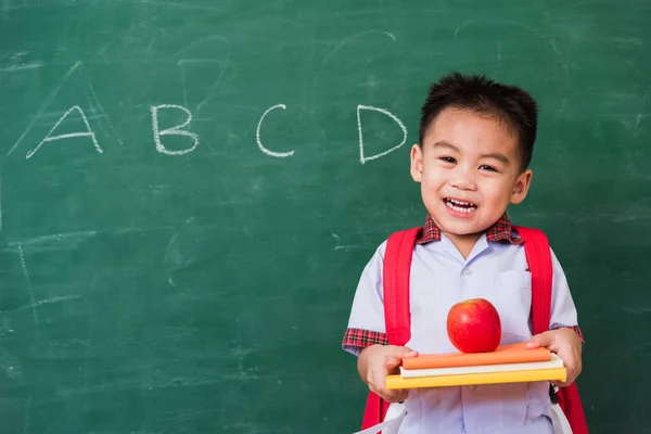 Back to School. Happy Asian funny cute little child boy from kindergarten in student uniform with school bag holding red apple on books smile on green school blackboard, First time to school education