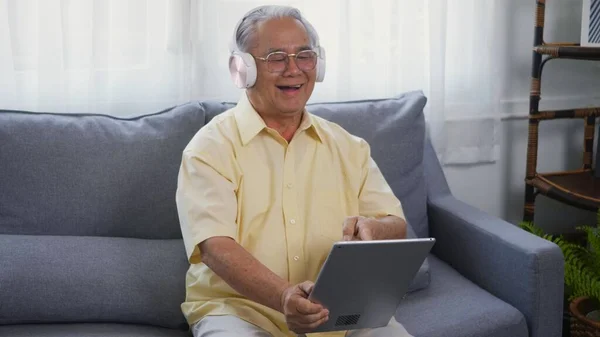 Old man grandfather smile with eyeglasses relaxing wear headphones is listening to music using a digital tablet, Asian happy senior old gray-haired sitting in living room on sofa at home video call