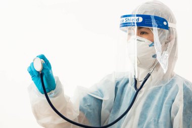 PPE üniformalı kadın tıp bilimci yüz maskesi ve plastik yüz kalkanı takarak Coronavirus 'u kontrol etmek için doktor stetoskobu (COVID-19) beyaz arka planda tutuyor.