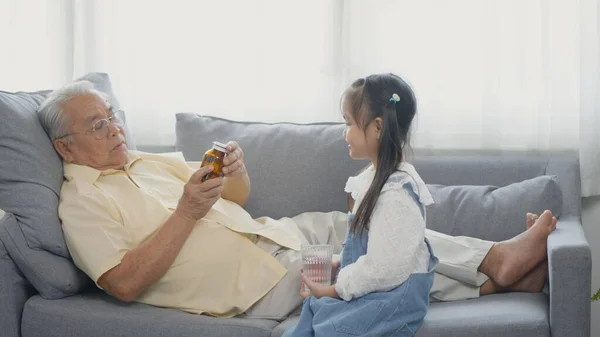 亚洲孙女带了药和水给爷爷在沙发上吃 老人在吃药的时候 孙女带着爱心照顾老人 — 图库照片