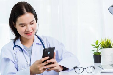 Asyalı doktor genç güzel bir kadın çalışırken veya akıllı cep telefonu ve dizüstü bilgisayarla hastane masasında gülümsüyor, teknolojik sağlık hizmetleri konsepti
