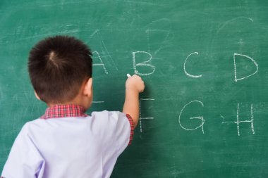 Okula geri dön. Küçük şirin çocuk anaokulunun mutlu Asyalı sırtı öğrenci üniforması içinde yeşil okul tahtasına beyaz tebeşirle ABC yazarak okul eğitimine ilk gelişi.