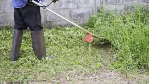 作業中の男性は 芝生のトリマー屋外で芝生を刈るブーツを着用 電気機械ガソリンブラシカッターを使用して切断草と男性のメンテナンス — ストック動画