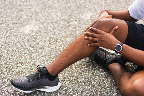参考译文 在室外街道健康公园跑步时 亚洲田径运动员黑色男子坐在手表上 用手抓住膝盖 这是他锻炼理念所造成的健康运动伤害 — 图库照片