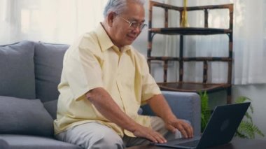 Kıdemli adam gözlük takıyor kanepeye oturuyor oturma odasında dizüstü bilgisayarda çalışıyor, mutlu yaşlı adam emekli oluyor, yaşlı büyükbaba evden çalışıyor, ağır çekimde çalışıyor.