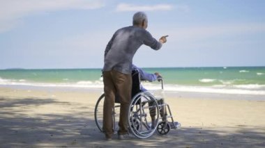 Mutlu Asyalı yaşlı kadın tekerlekli sandalyede oturuyor kocası da sahilde tekerlekli sandalye kullanıyor, yaz tatili, emekli çift konsepti.