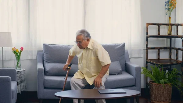 眼鏡をかけたおじいさんは 家の居間のソファからゆっくりと地面をきれいにするために杖を使っていた 膝の痛みに苦しむアジアのシニア古い白髪の痛み — ストック写真