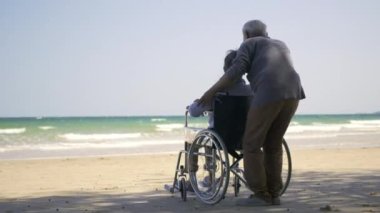 Mutlu Asyalı yaşlı kadın tekerlekli sandalyede oturuyor kocası da sahilde tekerlekli sandalye kullanıyor, yaz tatili, emekli çift konsepti.