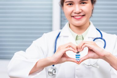 Kadın doktor gülümseyerek kalp şeklinde bir kalp oluşturuyor. Kalp parmağı, cam şişe koronavirüs aşısı, COVID-19 İlaç ve Sağlık Konsepti gösteriyor.