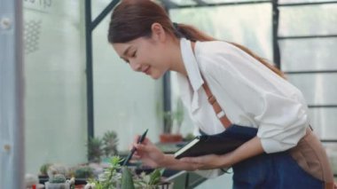 Asyalı güzel, genç bahçıvan kadın önlüğü ile kağıttan kitap kullanarak sera kaktüslerinin büyümesini kontrol ediyor. Çiçekçi ve küçük işletme konsepti var.