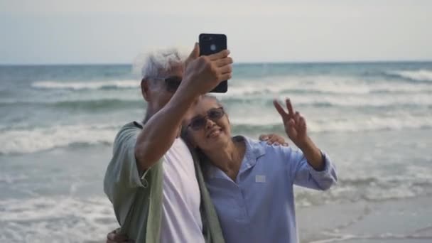 快乐的亚洲老年男女夫妇微笑着在海滩上用智能手机拍下自己的照片 浪漫的老年人享受暑假旅行 为退休夫妇的生活保险计划 — 图库视频影像