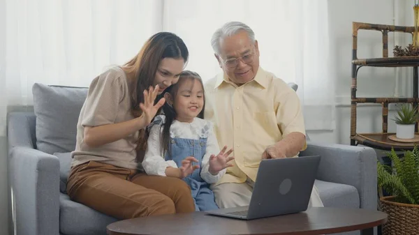 Happy family senior grandfather, daughter and granddaughter talking online via video chat using laptop computer with father, old grandpa embracing little grand child and mother in living room at home