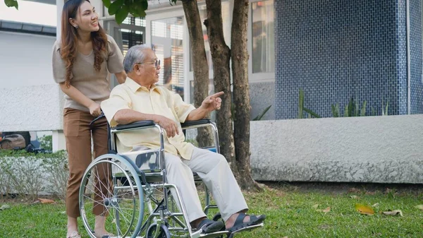 車椅子で身障者高齢者と娘 幸せなアジアの世代の家族が一緒に屋外の裏庭を楽しんで ケアヘルパー若い女性が笑顔で高齢者の男性を歩くと笑った — ストック写真