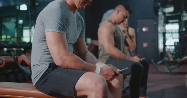 フィットネスクラブジムでトレーニングの練習後にスマートフォンを使用して座ってアクティブな若いグループ休憩 スポーツ健康的なライフスタイル技術の概念 — ストック動画