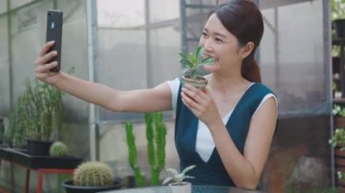 Mutlu Asyalı güzel genç bir kadın sera yakınlarındaki bir bahçede akıllı telefondan küçük bir ağacın selfie fotoğrafını çekiyor. Kadın bahçıvan videosu dijital cep telefonundan canlı yayında, ev bitkisinden.