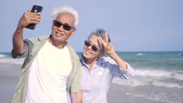 幸せなアジアのシニア男性と女性のカップルの笑顔ビーチでスマートフォンで自撮り写真を撮る ロマンチックな高齢者が楽しむ旅行夏休み 退職カップルのコンセプトで生命保険を計画 — ストック動画