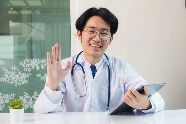 幸せなアジアの若い医師ハンサムな男性の笑顔で制服を着た眼鏡を手にして 患者を迎えるために手を上げます現代のスマートデジタルタブレットコンピュータ病院の机の上に テクノロジーヘルスケアと医学 — ストック写真