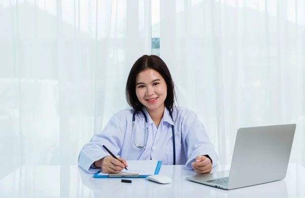 アジアの医師の若い美しい女性は ノートパソコンで作業を使用して笑顔と彼女は病院のデスクオフィスで書類やクリップボードの白い紙に何かを書く 医療の概念 — ストック写真