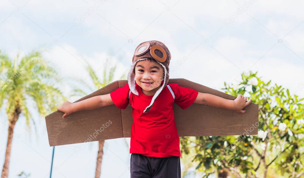 Happy Asian funny child or kid little boy smile wear pilot hat and goggles play toy cardboard airplane wing flying against summer sky cloud on trees garden background, Startup freedom concept
