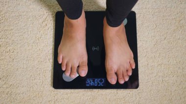 Asyalı genç bir kadın elektrik terazisine basıyor, kadın oturma odasında çalışıyor, kadın dijital tartı makinesinde duruyor. Sağlıklı kilo verme konsepti, yavaş çekim
