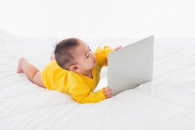 Asyalı küçük bir kız bebek dizüstü bilgisayarda elleri yazan sarı bir elbise giyiyor ve beyaz yataktaki monitöre bakıyor. Küçük bilişim adamı.