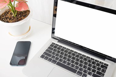 Modern çalışma alanı, dizüstü bilgisayarı boş beyaz ekran, gözlükler ve akıllı telefon beyaz masası masanın üzerinde saksılar, not defteri ve cep telefonu ev ofisinin üzerinde.
