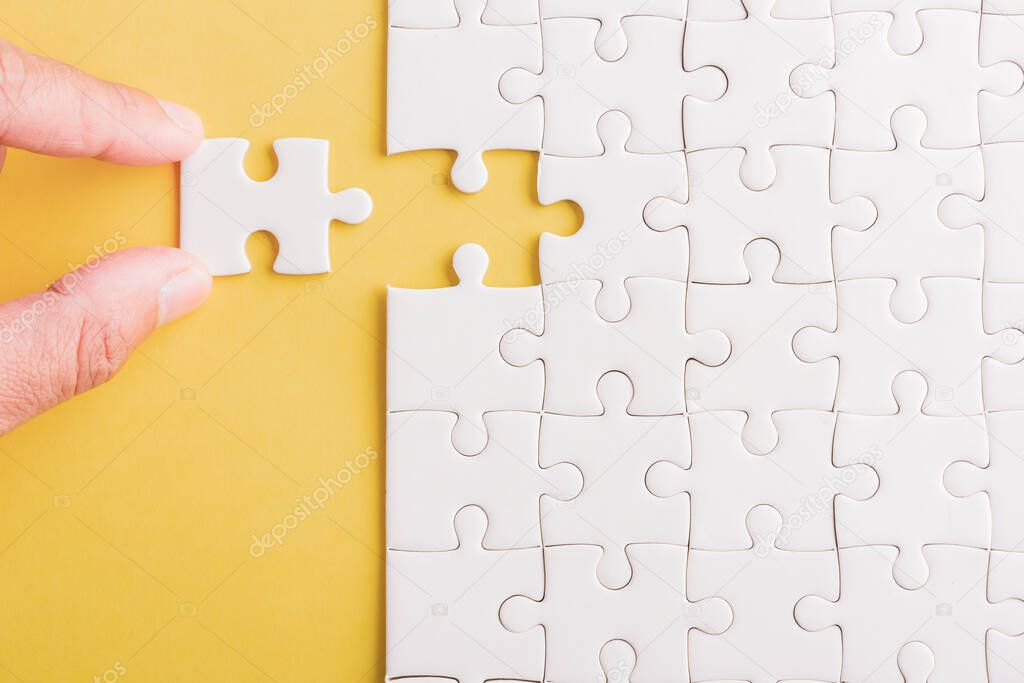 Top view flat lay of hand-holding last piece white paper jigsaw puzzle game last pieces put to place for solve problem complete mission, studio shot on a yellow background, quiz calculation concept