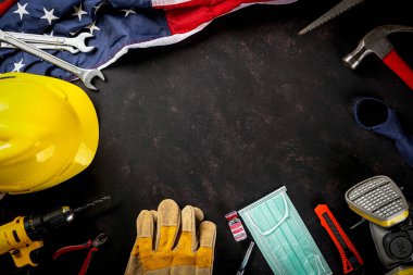 Mutlu İşçi Bayramı. Birçok mühendis çalışma araçları, tıbbi yüz maskesi koruyucu ve kopya alanı siyah arkaplanı olan Amerikan bayrağı, patlak koronavirüs konsepti sırasında Amerikan işgücü