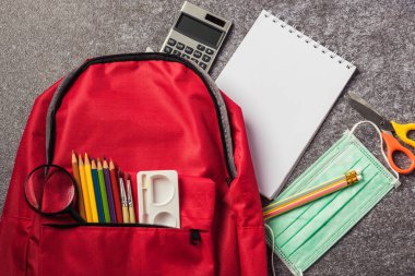 Masa başında yüz maskesi ve kırtasiye malzemeleriyle süslü kırmızı okul çantası, COVID-19 ya da Coronavirus konsepti salgını sırasında okula dönüş eğitimi.