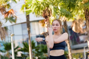 Formda Asyalı kadın dışarıda spor egzersizi yapıyor, kolunu esnetiyor, inanılmaz bir formda olduğunu gösteriyor, canlı ve sağlıklı insanları teşvik ediyor..