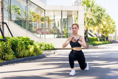 Formda bir kadın spor egzersizi yapar dışarıda hamleler yapar, sağlıklı ve aktif bir yaşam tarzı için bacaklarını açar, güneşli bir günde şehir parkına yakın bir yerde spor yapmak için kendini adadığını gösterir..
